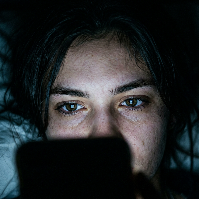 A cinematic close up of a teenager sitting in a dark room illuminated only by their smartphone screen